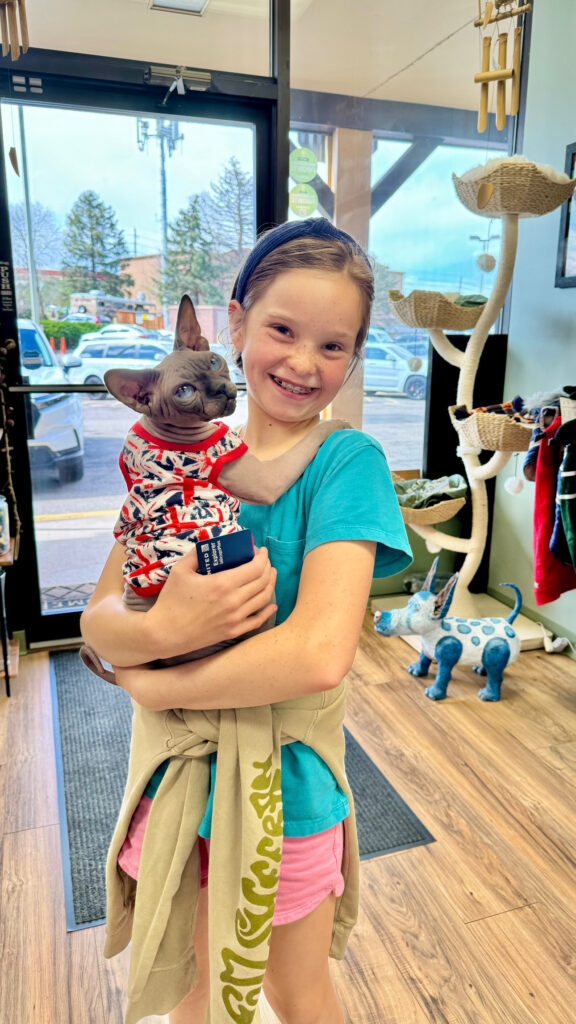 Smiling girl holds a Sphynx cat in patterned clothes inside a pet store with cat furniture and decor in the background.