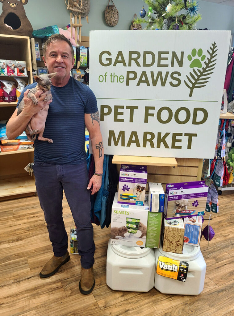 Man holding a small dog stands beside a "Garden of the Paws Pet Food Market" sign inside a pet supply store.