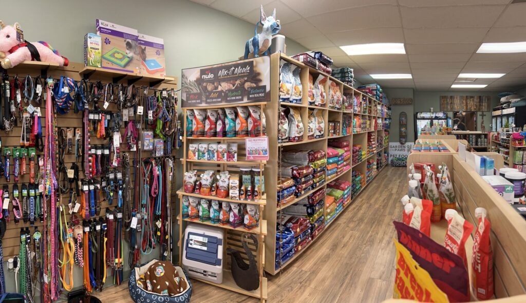 Pet supply store aisle with shelves of pet food, toys, collars, leashes, and various pet care products on display.